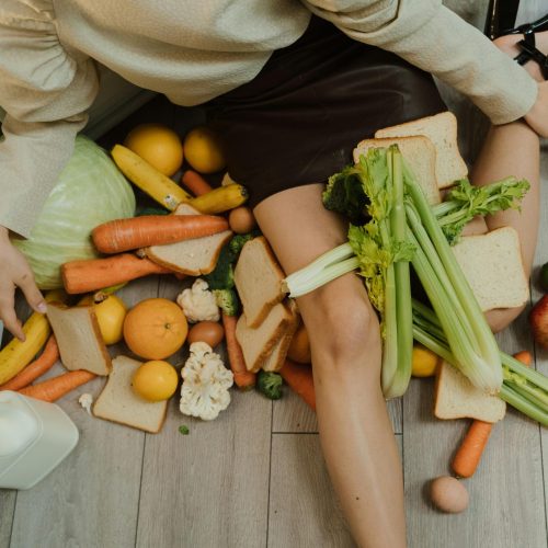 A woman amidst spilled groceries, highlighting food waste and consumerism with vegetables, fruits, and bread.